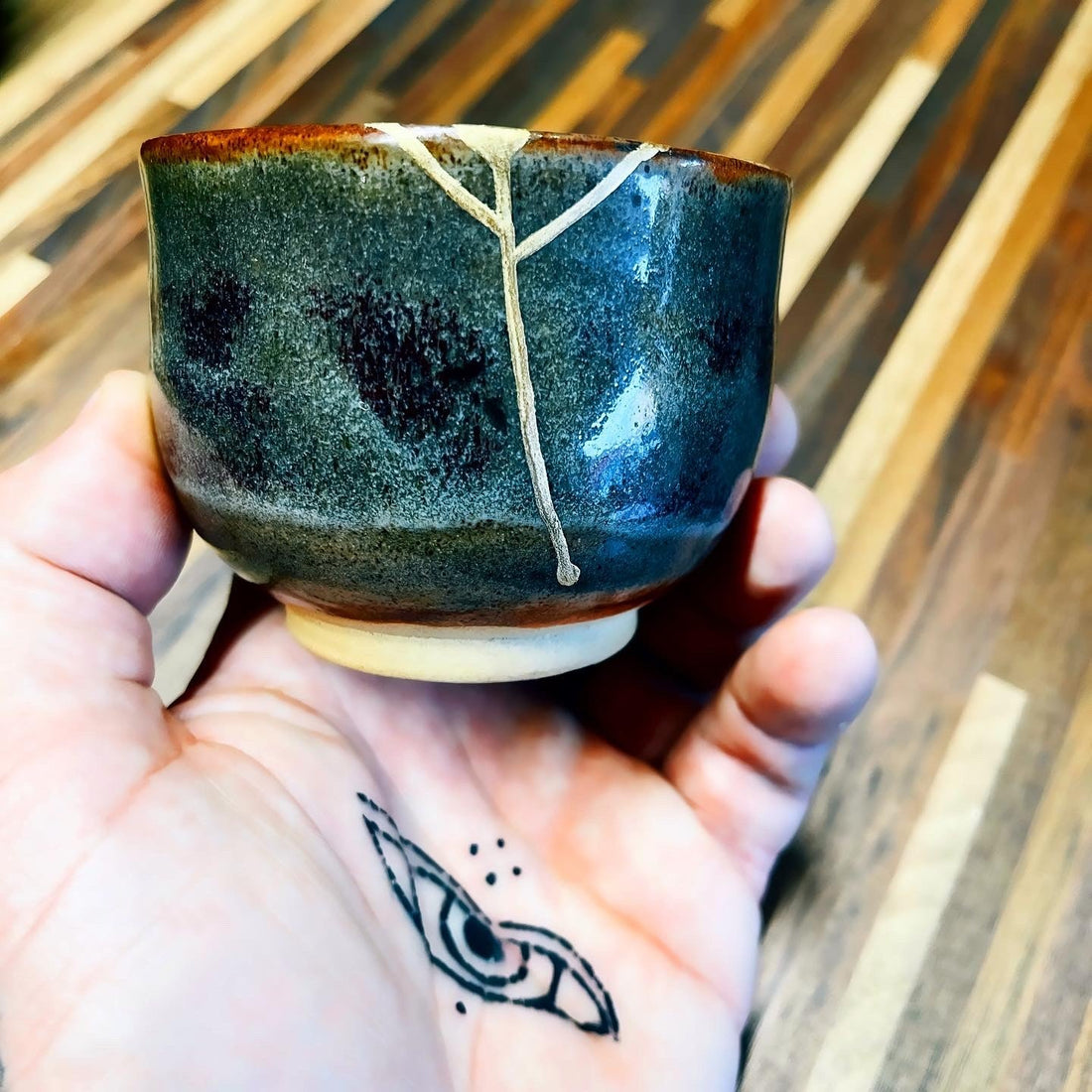 Petite tasse à glaçure vert sombre restaurée au kintsugi, ligne de réparation partant du bord, tenue au creux de la main
