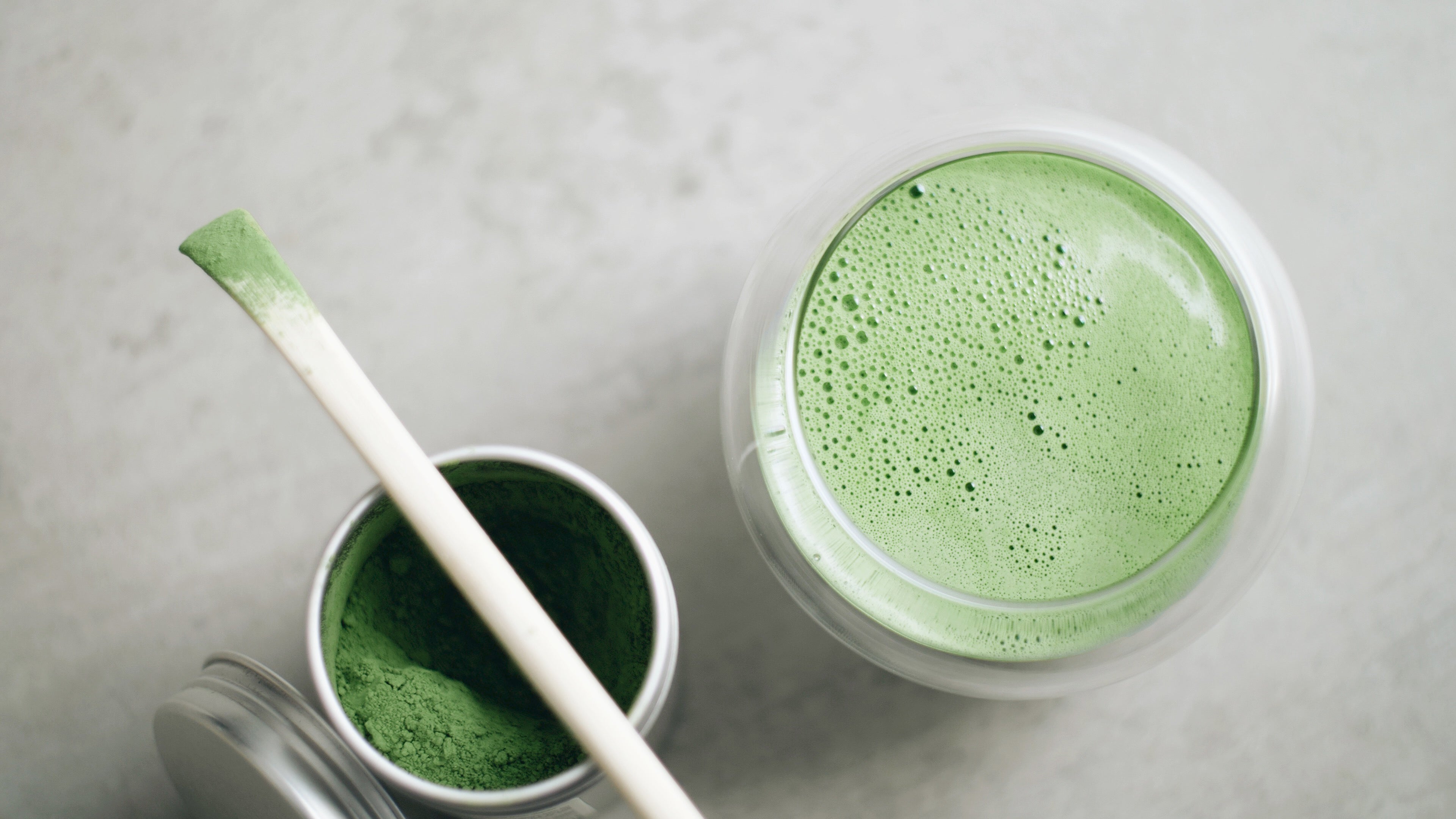 Un bol de matcha frais à côté d’un contenant de poudre de matcha vert avec une spatule en bambou, sur un fond neutre